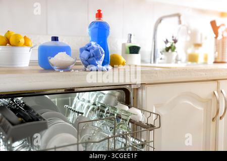 Dettaglio di una lavastoviglie incorporata in un armadietto della cucina con un piano di lavoro pieno di prodotti per lavare e mantenere la lavastoviglie automatica. Vista frontale. Foto Stock