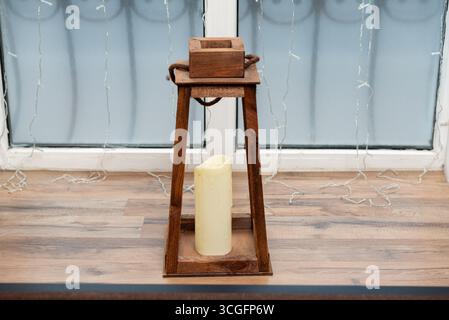 Un'affascinante lanterna rustica in legno caratterizzata da un'alta candela all'interno. sullo sfondo di finestre e luci fatate, questa scena crea un'intima inviti Foto Stock