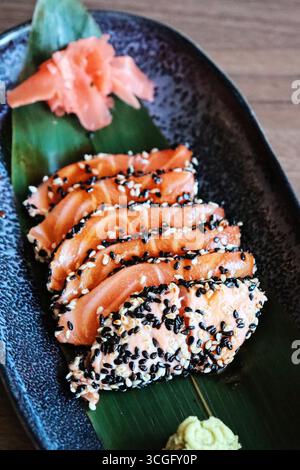 Un primo piano di sashimi di salmone fresco ricoperto di semi di sesamo bianco e nero, servito su una foglia verde con zenzero sottaceto e wasabi su un piatto scuro. Foto Stock