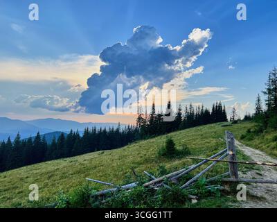 La spettacolare formazione di nuvole domina il cielo serale sul tranquillo paesaggio montano. Alti pini e colline erbose sono in primo piano, con recinzione rustica in legno e sentiero che conduce alla scena. Foto Stock