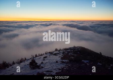 La splendida fotografia del paesaggio cattura la bellezza serena della cima innevata della montagna all'alba, con un mare mozzafiato di nuvole che si estende all'orizzonte. Il cielo passa da blu intenso a arancione caldo. Foto Stock