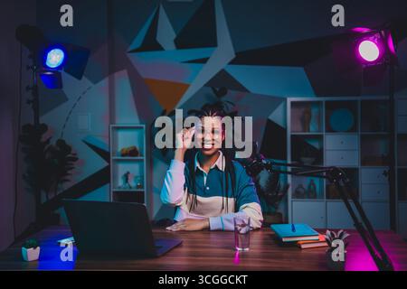 Donna che ha trasmesso la radio per registrare un podcast in un elegante ufficio domestico con luci al neon in un ambiente creativo Foto Stock