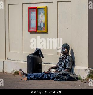 Un artista di strada di Brighton che suona un banjo e si veste di nero appoggiato su un muro di crema. Foto Stock