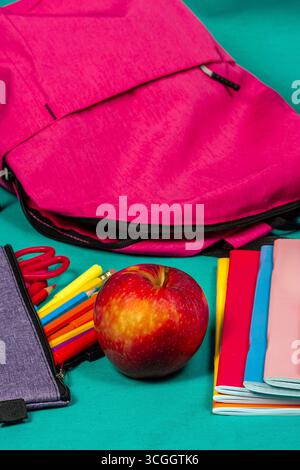 Zaino rosa, mela, astuccio e quaderni appoggiati su una superficie turchese Foto Stock