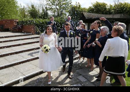 Una coppia appena sposata che cammina lungo una serie di gradini di pietra all'aperto, tenendosi per mano e sorridendo mentre vengono bagnate di coriandoli da familiari e amici Foto Stock