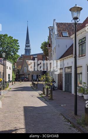 Una tranquilla e stretta strada acciottolata a Gouda fiancheggiata da tradizionali case olandesi, che conduce verso una lontana guglia della chiesa "De Gouwekerk" sotto una chiara b Foto Stock