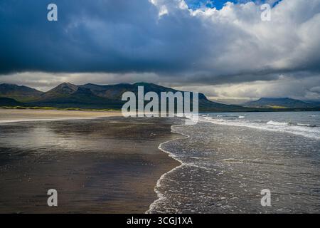 Le sabbie scure incontrano dolci onde sotto i cieli tempestosi in Islanda, dove le spettacolari montagne si innalzano oltre la costa in una scena di pura bellezza Foto Stock