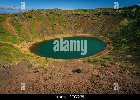 Un'ampia vista sul lago del cratere di Kerið in Islanda, che mostra le sue acque blu profonde incorniciate da ripidi pendii vulcanici di rosso e verde Foto Stock