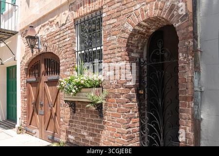 Charleston, South Carolina, Stati Uniti - 2 luglio 2025: Una storica parete in mattoni con una porta ad arco in legno, cancello in ferro battuto e una scatola di fiori piena di colon Foto Stock