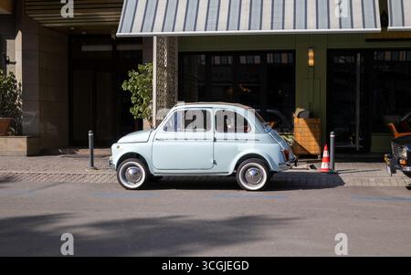 Vista laterale di una piccola Fiat 500 d'epoca azzurra parcheggiata su una strada soleggiata di fronte a un moderno bar o negozio Foto Stock