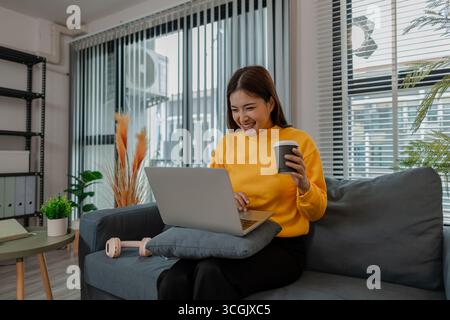 Giovane donna asiatica in felpa gialla sorridente mentre guarda qualcosa sul suo portatile e tiene una tazza di caffè. Si siede comodamente su un divano in una Foto Stock
