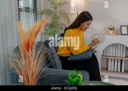Giovane donna asiatica in maglione giallo seduta sul divano e sorridente mentre scrive appunti sul taccuino. Atmosfera rilassata in un accogliente soggiorno. Concetto di le Foto Stock