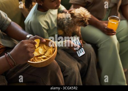 Bambini neri seduti con adulti neri sul divano, con il cane piccolo e il telecomando della TV, mangiando patatine fritte dalla ciotola, bevendo una bevanda all'arancia, guardando la televisione insieme Foto Stock