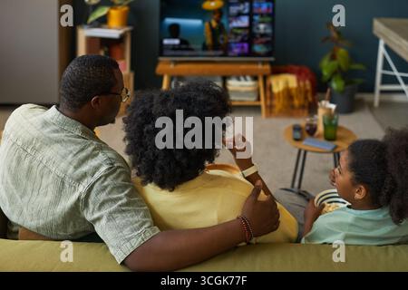 Uomo nero seduto con donna nera e bambino nero sul divano a guardare la televisione insieme, uomo che mette il braccio intorno alla donna, famiglia che trascorre il tempo libero a casa Foto Stock