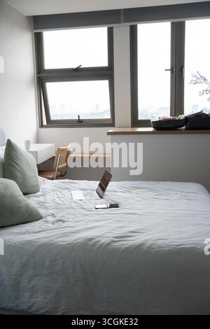 Computer portatile e smartphone aperti posizionati su un letto appena fatto in una camera d'hotel luminosa e confortevole, che suggerisce di lavorare a distanza durante i viaggi Foto Stock