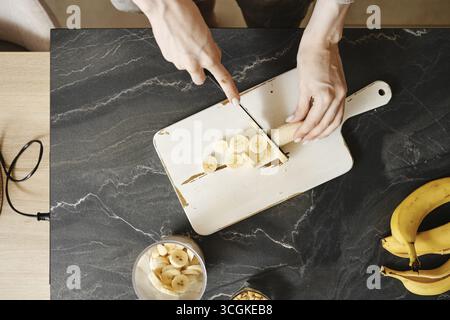 Una persona affetta banane fresche su un tagliere bianco in una cucina ben illuminata. Il piano di appoggio è dotato di una ciotola di banane e di un vaso di vetro. Esposizione alla luce solare Foto Stock