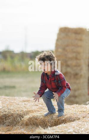 Bambino in camicia a quadri rossa accovacciato su balle di fieno all'aperto Foto Stock