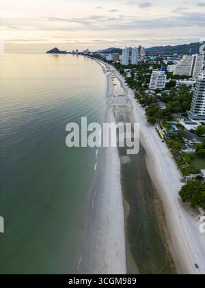 Vista aerea della tranquilla spiaggia che incontra lo skyline della città con edifici moderni e una vegetazione lussureggiante sotto un cielo sereno, Hua Hin, Prachuap Khiri Khan, Thailandia. Foto Stock