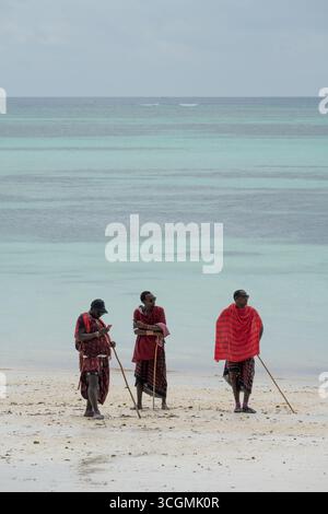 Tre uomini Maasai in abiti rossi tradizionali in piedi sulla spiaggia, uno con un telefono cellulare, che mostrano la tradizione incontra la vita moderna. Foto Stock