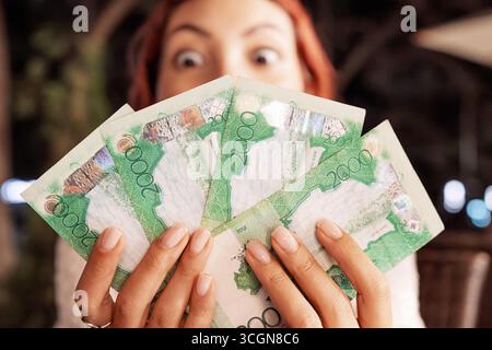 Una giovane donna sorpresa che tiene un fan delle banconote in stile kazako davanti al suo viso, espressione dagli occhi larghi Foto Stock