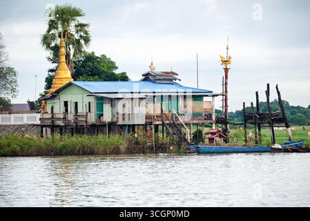 Ayeyarwaddy River Stilt House con Golden Pagoda e Hintha Bird Mandalay Myanmar // MANDALAY, Myanmar — Una tradizionale palafitta si trova sulle rive del fiume Ayeyarwady (noto anche come Irrawaddy River o Ayeyarwady River) a Mandalay. La scena presenta una pagoda dorata (stupa) e un'importante scultura dorata dell'uccello Hintha (noto anche come uccello Hamsa), che è un simbolo significativo nella cultura birmana, spesso rappresentando la purezza e la prosperità. Le palafitte si trovano comunemente lungo il fiume Ayeyarwady, fornendo case per le comunità adattate ai livelli fluttuanti dell'acqua del fiume. Questi Foto Stock