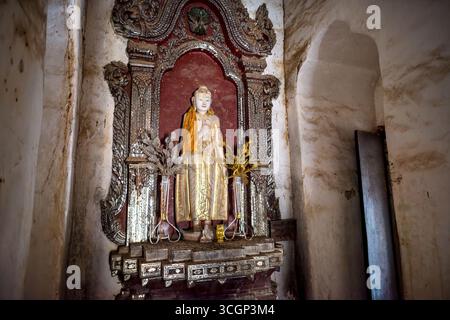 Monastero di Maha Aung Mye Bon San Statua di Buddha ornata e intarsiata Niche Amarapura Myanmar // AMARAPURA, Myanmar — Una statua di Buddha in piedi è custodita in una nicchia ornata presso il monastero di Maha Aung Mye Bon San (noto anche come me Nu Oak Kyaung) ad Amarapura, regione di Sagaing. La statua raffigura il Buddha con il mudra Abhaya (mano destra sollevata) e il mudra Varada (mano sinistra abbassata), simboleggiando l'impavidità e la generosità. Caratteristica della tradizionale arte buddista birmana, l'intricata nicchia è adornata con dettagliate incisioni e intarsi a specchio o madreperla. La monasta di mattoni del XIX secolo Foto Stock