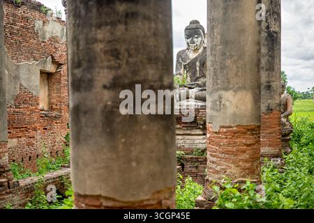 Complesso della Pagoda di Yadana Hsemee, Statua di Buddha, rovine di mattoni Inwa Myanmar // INWA, MYANMAR — Una statua di Buddha intemprata in posizione seduta è visibile tra le rovine di mattoni e la vegetazione ricoperta del complesso della Pagoda Yadana Hsemee. Questo antico sito fa parte di Inwa (conosciuta anche come Ava), un'ex capitale reale dei regni birmani. Il complesso presenta numerose pagode in mattoni e immagini di Buddha, spesso realizzate in stucco su mattoni. Queste strutture hanno gravi danni da secoli di esposizione e da terremoti significativi, tra cui il tremore del 1839. Situato vicino ad Amarapura, nella Sagain del Myanmar Foto Stock