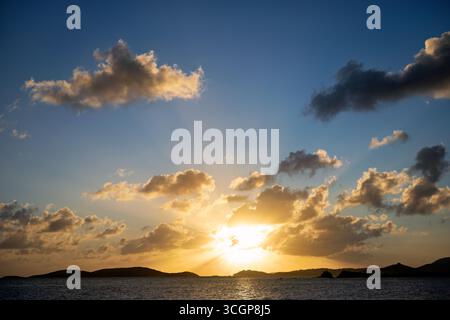 Tramonto su San Tommaso da Gallows Point Cruz Bay St John USVI // ST JOHN USVI - Un tramonto illumina il cielo con raggi dorati che si infrangono attraverso le nuvole sull'isola di St. Thomas in lontananza. Questa vista viene catturata da Gallows Point a Cruz Bay, St. John, parte delle Isole Vergini americane. Le tranquille acque del Mar dei Caraibi riflettono le calde sfumature del sole che tramonta. St. John è conosciuta per le sue spiagge incontaminate e la lussureggiante bellezza naturale, che fanno parte dell'arcipelago delle Isole Vergini. Foto Stock