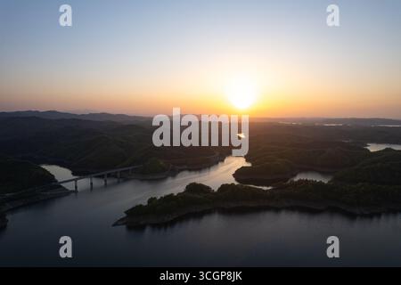 Lo splendido tramonto dipinge il cielo sopra il Danjiangkou Reservoir, Cina, illuminando lussureggianti colline verdi e acque tranquille con un bagliore dorato. Foto Stock