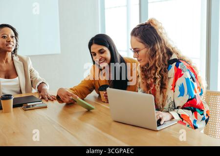 Gruppo di colleghi che collaborano durante una riunione di lavoro. Tre donne condividono idee mentre utilizzano un computer portatile e uno smartphone. L'ambiente d'ufficio Foto Stock