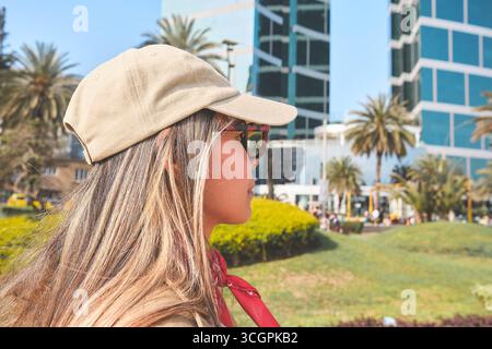 Giovane donna che indossa occhiali da sole e berretto beige per esplorare il parco della città moderna con palme e grattacieli Foto Stock