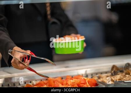 Primo piano delle mani di una donna con lunghe unghie rosse che usano pinze metalliche per servire il cibo in una caffetteria a pagamento. Indossa una giacca nera. Foto Stock