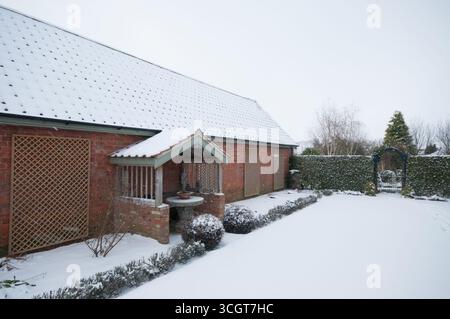 Una casa in mattoni con tetto innevato si trova vicino ad un bellissimo giardino caratterizzato da un pozzo, circondato da una siepe in un tranquillo paesaggio invernale. Foto Stock