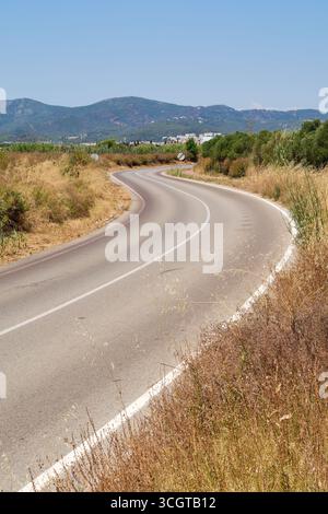 Una tortuosa strada asfaltata serpente attraverso un paesaggio rurale estivo. In lontananza, le montagne e una città sono visibili sotto un cielo azzurro. Foto Stock