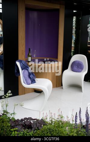 Due eleganti sedie bianche con cuscini viola si trovano in un giardino contemporaneo, caratterizzato da piante decorative e un elegante allestimento per vini. Foto Stock