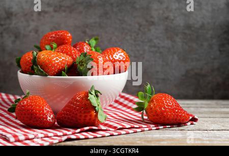 Concetto di fragola con un gruppo di fragole rosse mature in una ciotola bianca primo piano vista frontale su un tavolo in legno rustico d'epoca e sfondo grigio. Foto Stock