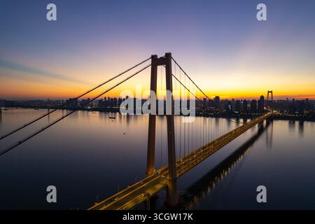 Un maestoso ponte attraversa un ampio fiume mentre il sole tramonta sul paesaggio urbano, illuminando il cielo. Foto Stock