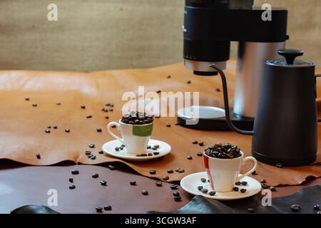 Concetto di stile di vita del caffè e della pelle: Tazze da caffè ripiene di chicchi di caffè su superfici artigianali in pelle, con macchina per il caffè e bollitore. Un creativo Foto Stock