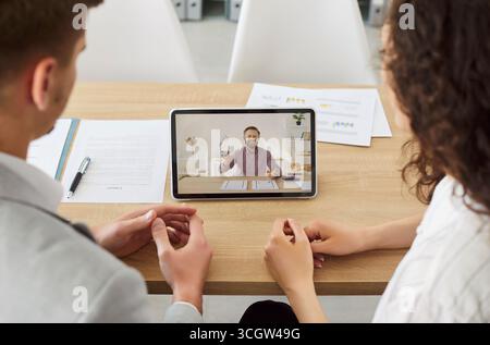 Gli uomini d'affari seduti in ufficio hanno una videochiamata online con un collega di sesso maschile che usa un tablet. Foto Stock