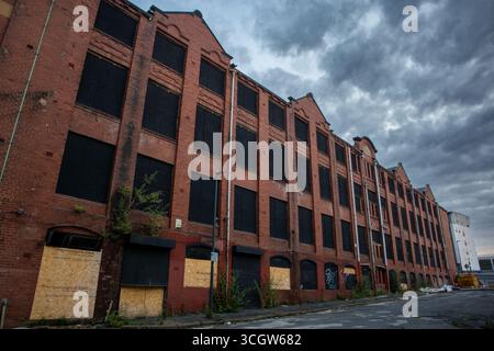 Grande magazzino di mattoni abbandonati con file di finestre a bordo, erbacce sovrastate e segni di decadimento, simboleggiando il declino e l'abbandono urbano Foto Stock