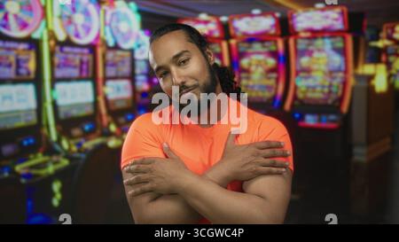 Uomo sorridente con le braccia incrociate sul petto e le mani sulle spalle accanto alla slot machine nell'edificio del casinò; tranquillità. Foto Stock