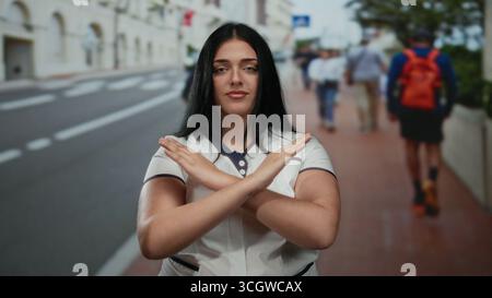 Donna sicura di sé con le braccia incrociate si staglia sulla strada della città, incarnando potenza e sfida in un ambiente urbano all'aperto con piedistallo sfocato Foto Stock