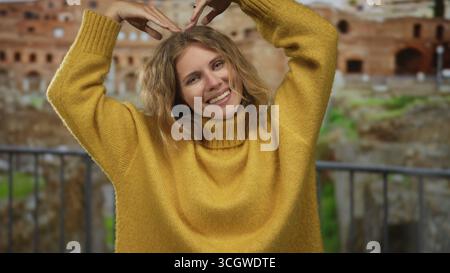Una donna solleva le braccia a forma di cuore vicino al panorama di roma mentre un giovane turista biondo emette un sorriso caldo indossando un comodo maglione giallo mentre ammira il paesaggio Foto Stock