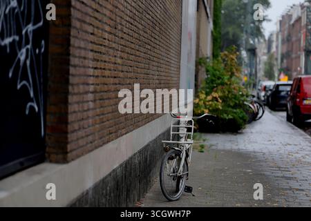 Bicicletta bianca parcheggiata contro un muro di mattoni su un marciapiede bagnato sotto la pioggia, Amsterdam, Paesi Bassi, messa a fuoco poco profonda che mostra la strada lontana e le auto. Foto Stock