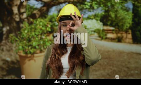 Una donna che indossa un elmetto fa un buon gesto nel parco soleggiato, mescolando lavoro e svago come architetto o ingegnere in un ambiente all'aperto vivace, incarnando pr Foto Stock
