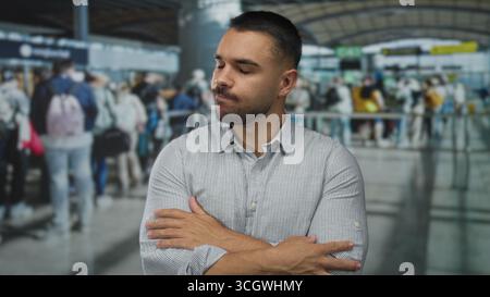 Uomo ispanico in camicia a righe con bottoni chiude gli occhi e si incrocia le braccia in mezzo ai viaggiatori nell'affollato terminal aeroportuale; negazione. Foto Stock