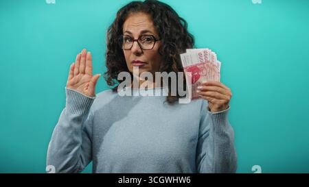 Donna di mezza età che tiene in mano banconote della corona islandese mentre alza la mano con un gesto di giuramento su uno sfondo blu. Foto Stock
