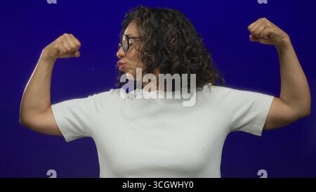 Donna ispanica di mezza età che flessa con sicurezza i muscoli contro un muro isolato di sfondo blu, mostrando forza e potenza nel suo casual Foto Stock