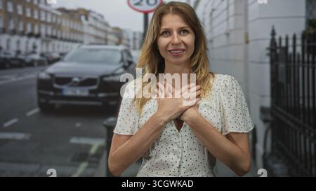 Una donna con un abito bianco a pois mette le mani sul petto in un gesto grato su una strada della città con auto parcheggiata e un cartello visibile a 20 mph; gratitudine heali Foto Stock