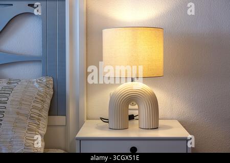Primo piano di un'elegante lampada da tavolo color crema con paralume in tessuto su un moderno comodino accanto a un cuscino a motivi geometrici. Foto Stock